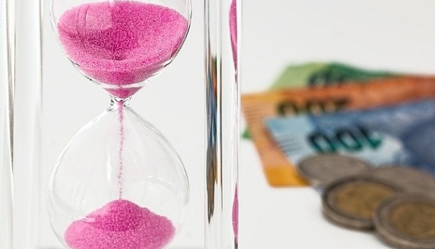 Das Bild zeigt eine Sand Uhr mit lila Sand und Geld im Hintergrund